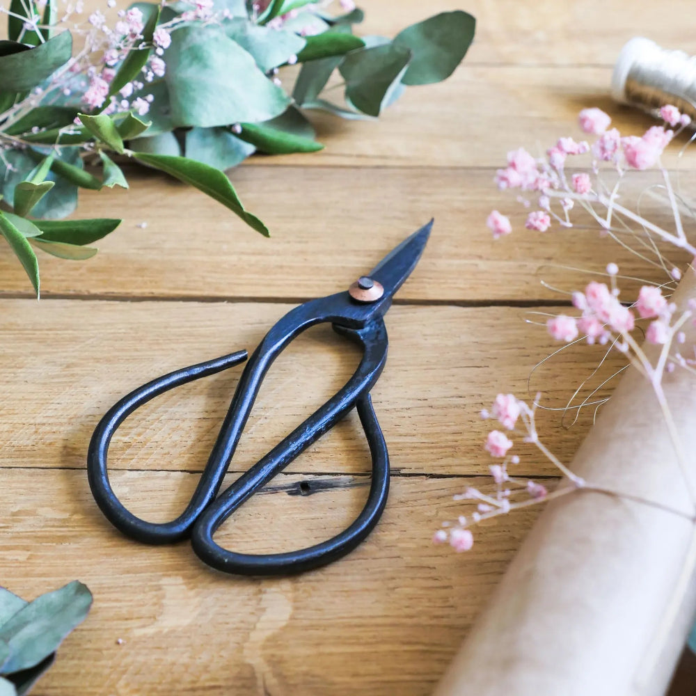 Japanese-style pruning shears on a wooden table, surrounded by greenery and dried flowers – perfect for indoor plant care and bonsai trimming.
