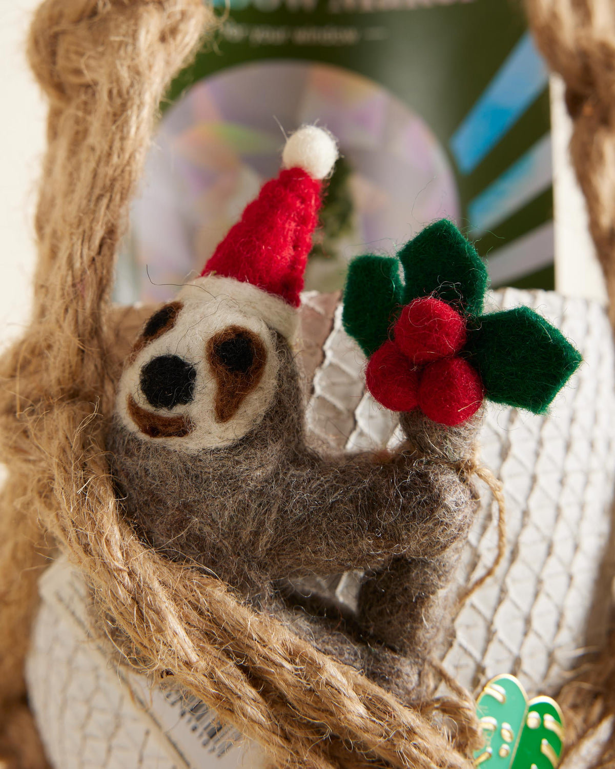 Felted sloth ornament wearing a Santa hat with holly leaves and berries on a textured background.