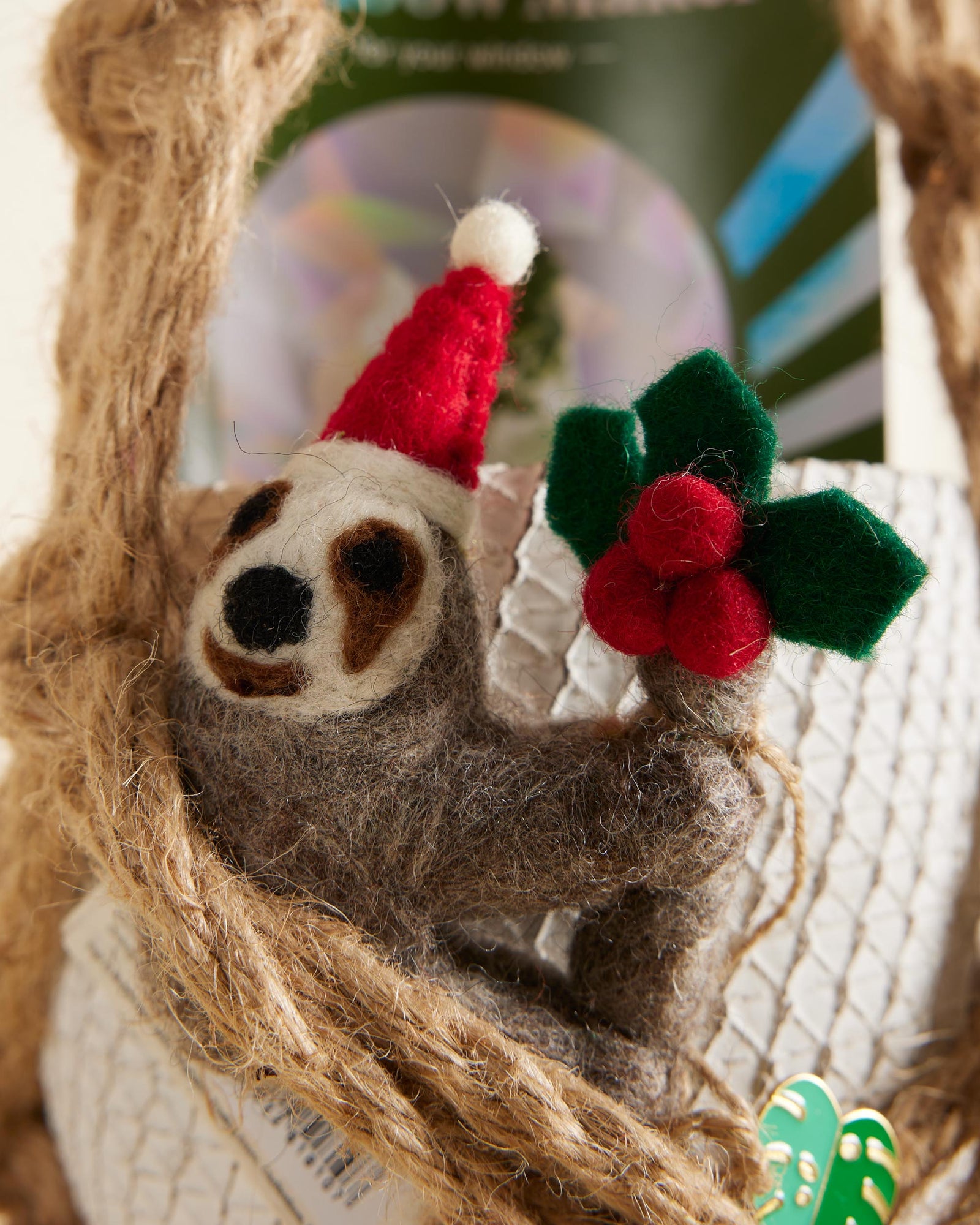 Felted sloth ornament wearing a Santa hat with holly leaves and berries on a textured background.